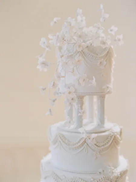 Wedding cake with cascading white sugar flowers and ornate piped frosting garlands, pearl borders, and tiers against a neutral backdrop