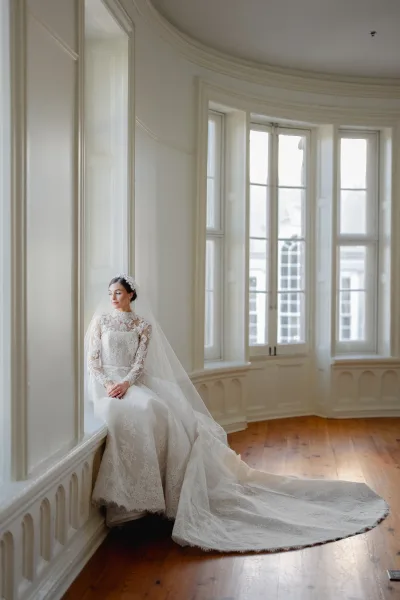 Bridal portrait of a bride in a lace wedding dress with long sleeves and cathedral veil, seated by tall windows in a white paneled room
