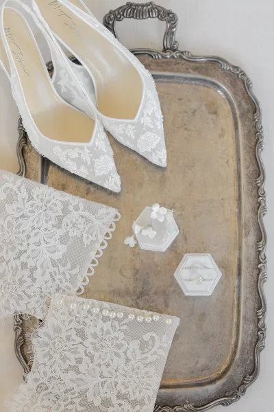 Bridal accessories flat lay featuring white lace wedding shoes, lace gloves, pearl bracelet, and engagement ring in a velvet box on light fabric
