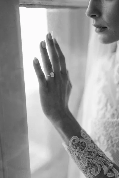 Engagement ring with an oval diamond sparkling on a bride’s manicured hand, framed by a lace sleeve in soft window light indoors