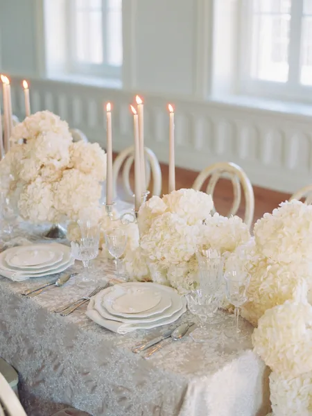 Reception tablescape with white hydrangea centerpiece, taper candles, silver candelabra, and crystal stemware in soft window light on wood floor