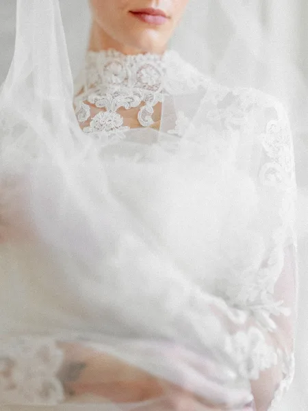 Bridal portrait in a lace wedding dress with high neck lace and a sheer veil over her face, posed against a neutral wall
