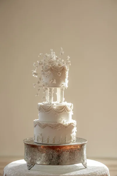 Wedding cake three tier wedding cake with piped frosting garlands and sugar flowers on an ornate silver stand against a beige backdrop