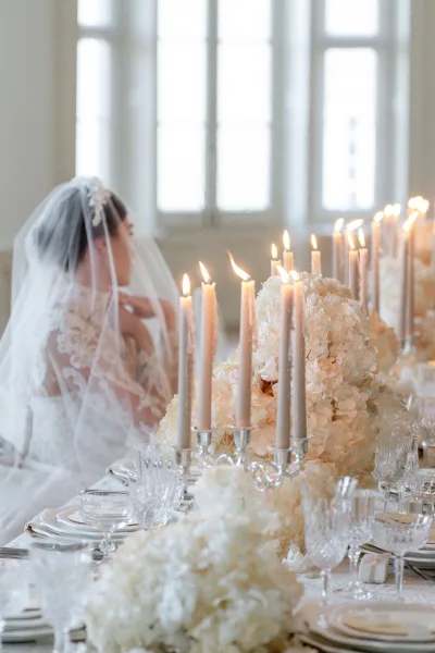 Wedding tablescape with taper candles in silver candelabras, white floral centerpieces and crystal glassware beside a bride’s lace veil by large windows