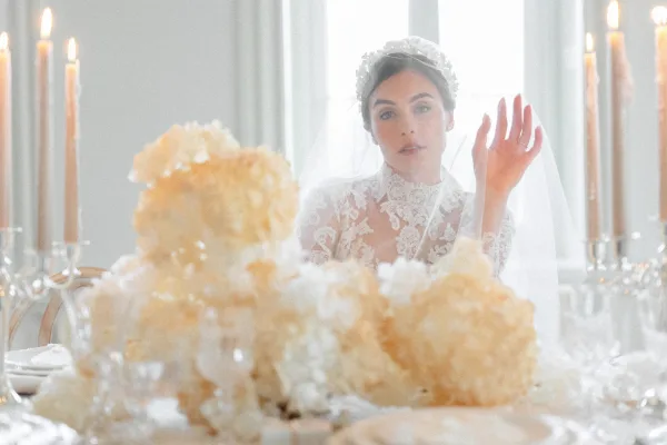 Bridal portrait of a bride in a lace high neck wedding dress and veil seated at an all-white reception table by large windows