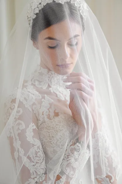 Bridal portrait of a bride under veil in a lace wedding dress, looking down and showing her engagement ring against a light neutral wall