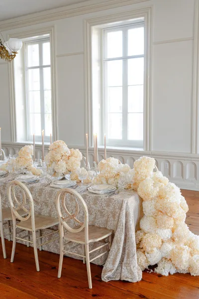 Reception tablescape with wedding head table decor featuring white florals, taper candles, crystal glassware, and chandelier-lit room with bright windows