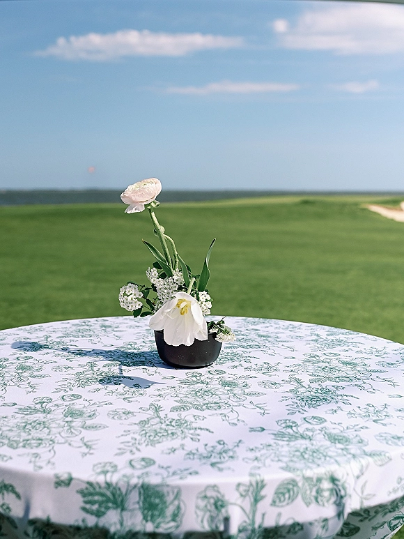 Wedding centerpiece with a small white floral arrangement in a black vase on a patterned tablecloth, set against a green lawn and blue sky