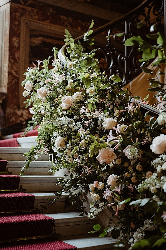 Staircase floral decor with a cascading garland of blush roses and greenery along a wrought iron railing on marble steps with red runner