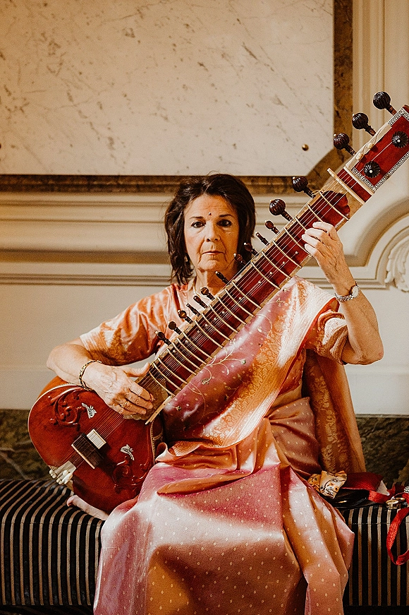 Wedding musician portrait of a sitar player in a silk sari with bindi, seated on a striped bench against a marble wall with ornate molding
