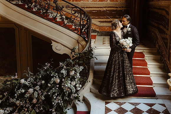 Wedding couple portrait of bride and groom on staircase sharing a kiss, she holds a bouquet as he wears a black tuxedo on a grand marble stairway