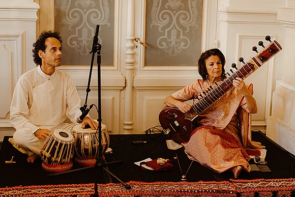 Wedding musicians in traditional Indian outfits perform live wedding music with tabla drums on rugs before ornate wall paneling indoors