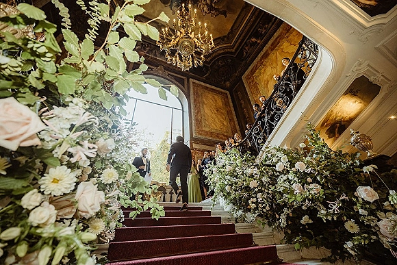 Ceremony processional as the groom walking down aisle on a red carpet runner past wedding aisle flowers in an ornate grand staircase foyer