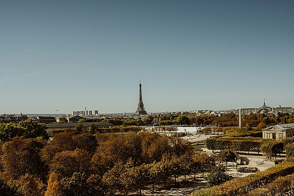 City skyline with autumn cityscape over golden treetops, park paths and rooftops under a clear blue sky with a distant tower
