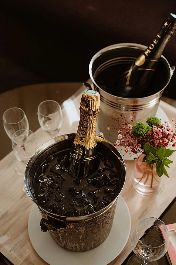 Champagne bucket with champagne on ice, bottle and glasses on a wood table, with a small floral arrangement nearby