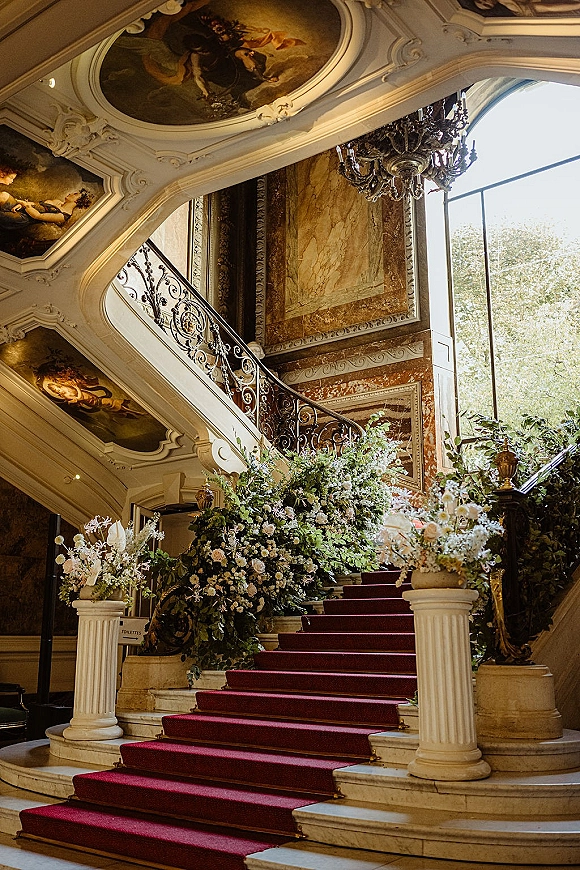 Grand staircase decor with wedding staircase flowers, lush greenery on railings, a red carpet runner, and chandelier in a marble hall