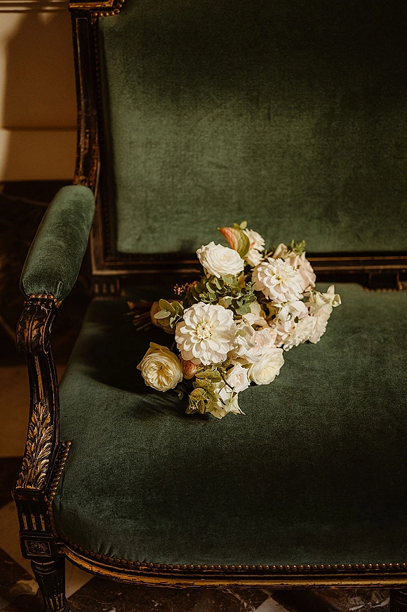 Bridal bouquet of white wedding bouquet blooms with lush greenery and ribbon draped over a vintage velvet chair on marble floor indoors