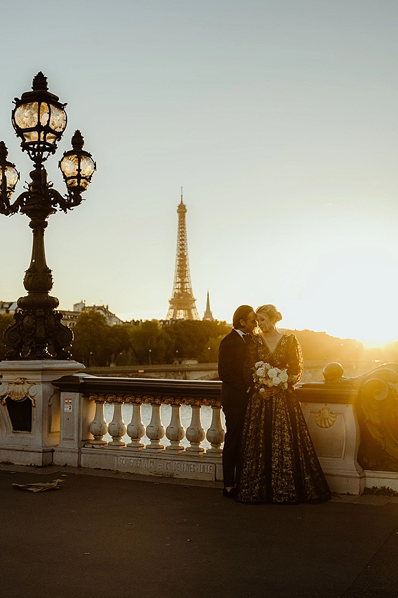 Couple portrait at sunset with bride holding a bouquet beside groom in black suit, leaning on bridge railing by river and skyline
