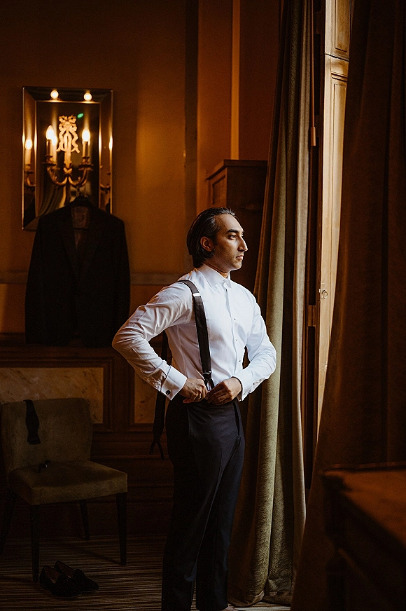 Groom getting ready by a hotel window, adjusting suspenders over a white dress shirt with cufflinks in warm sconce light