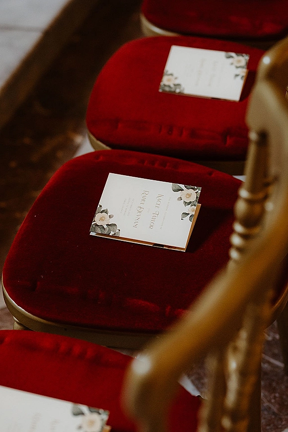 Ceremony programs with botanical floral stationery rest on gold chairs with velvet cushions in an aisle seating row on the floor