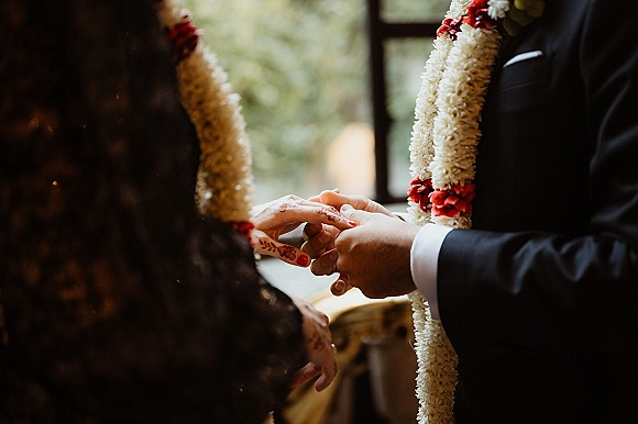 Wedding ring exchange as groom slides band onto bride’s hennaed hand with red nail polish, floral garlands visible near a window-lit ceremony space