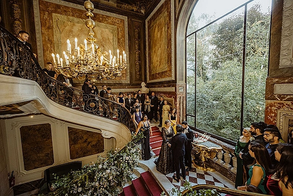Wedding guests on stairs in formal attire on a grand staircase, gathered by a red carpet runner beneath a chandelier in a marble foyer