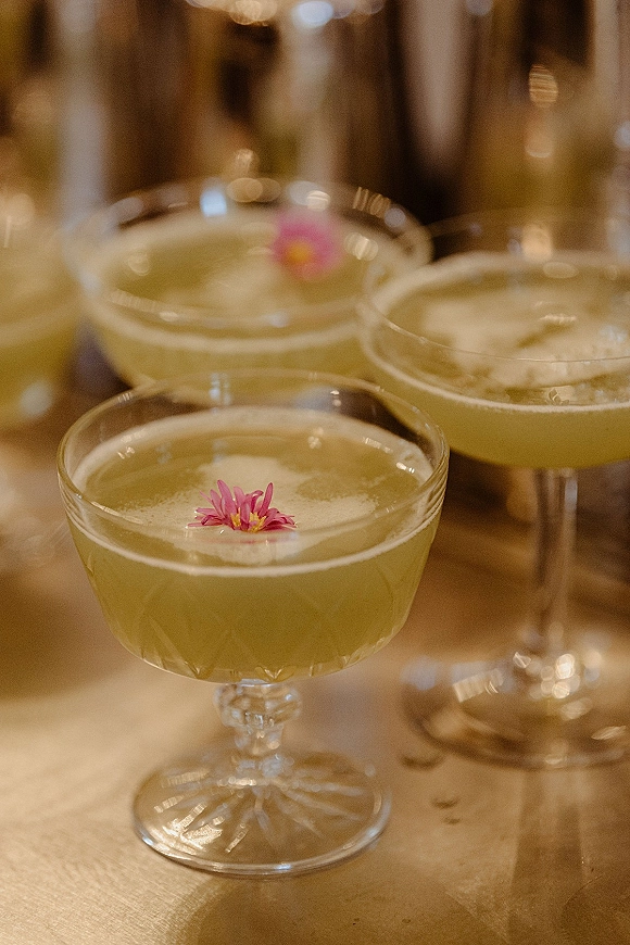 Signature cocktails topped with cocktail foam in coupe glasses, garnished with pink edible flowers on a gold bar with warm lights