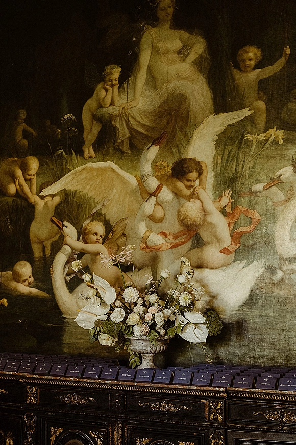 Wedding escort cards in navy displayed alphabetically on a ledge above a vintage sideboard, with an urn of white blooms and greenery against an ornate mural backdrop