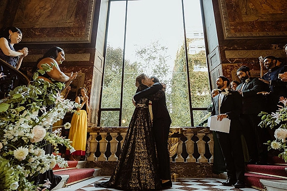 Wedding kiss as the couple embraces on a grand staircase, bride in black lace gown holding a bouquet, guests cheering by a tall window