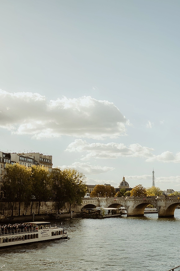 City river view with a stone bridge over river and a river cruise boat passing, framed by city buildings, trees, and cloudy sky