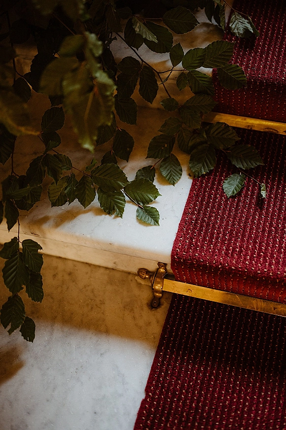 Wedding flat lay with greenery garland and burgundy woven runner styled along marble steps beside a brass railing