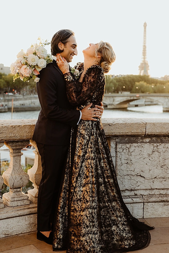 Couple portrait with a wedding bouquet portrait as bride touches groom’s face by a stone balustrade, river, bridge, and sunset skyline