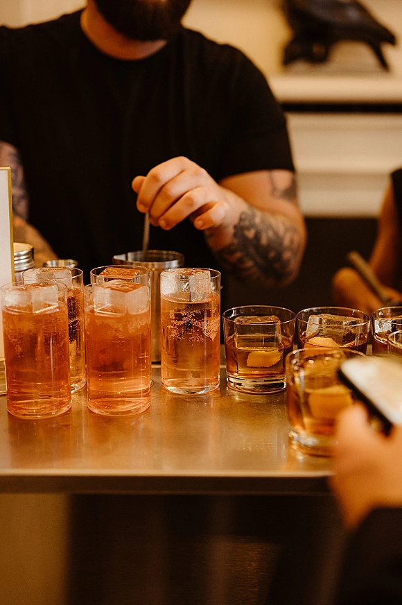 Wedding cocktails lined on a bar counter, signature wedding drinks on ice in glassware with napkins and a small table sign, bartender behind
