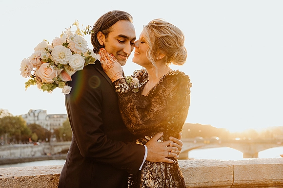 Couple portrait at sunset, couple embracing wedding with foreheads touching as bride cups groom’s face, river and stone bridge behind