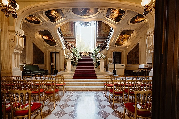 Ceremony setup for an indoor wedding ceremony with red aisle runner, gold Chiavari chairs, chandeliers, and florals by a grand staircase