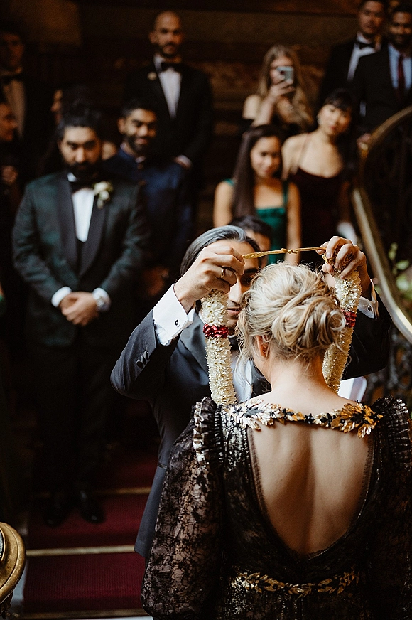 Ceremony moment as bride and groom exchange a flower garland, tuxedo boutonniere and guest attire visible on an indoor staircase