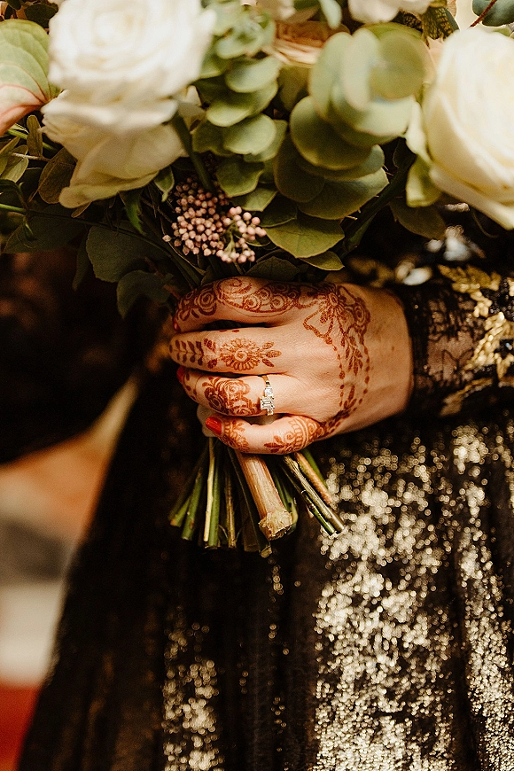 Bridal bouquet close-up of a white rose bouquet with eucalyptus, held in hennaed hands with an engagement ring and red nails indoors
