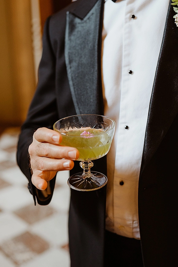 Wedding cocktail in a coupe glass, green signature wedding drink with edible flower garnish held by a groom in tuxedo under warm indoor lighting