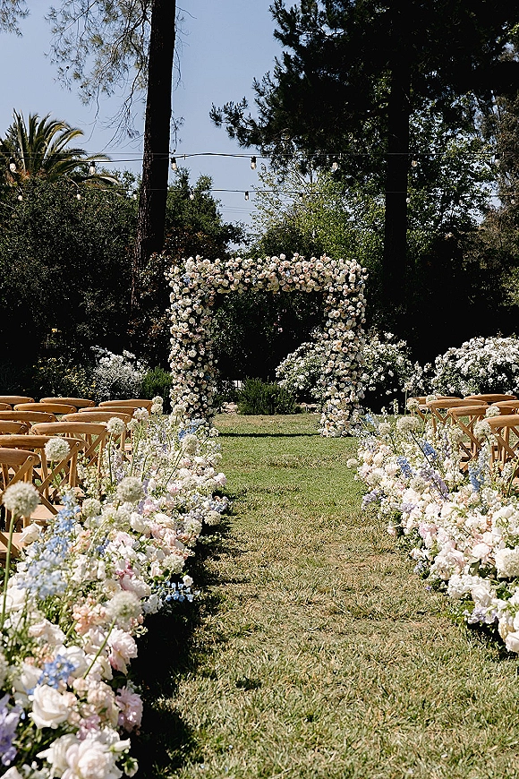 Ceremony aisle decor with pastel rose aisle flowers leading to a floral arch, wooden crossback chairs under string lights on a grassy lawn