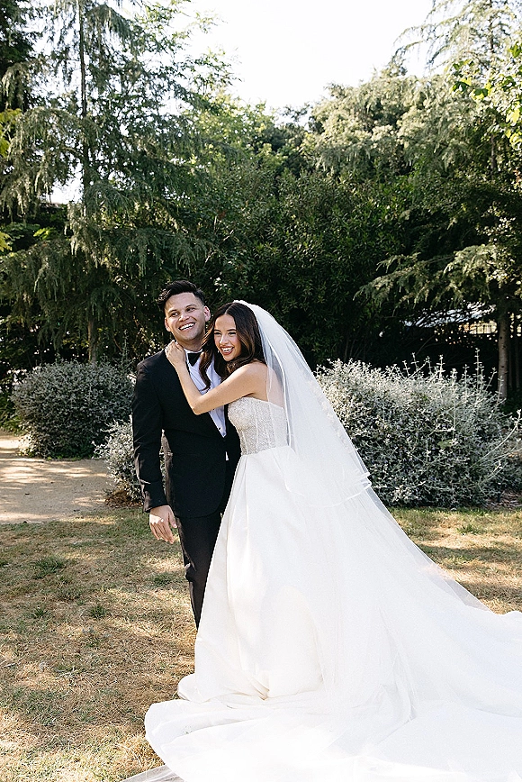 Couple portrait of bride hugging groom in a strapless gown and long veil, with groom in tuxedo, on a sunlit garden lawn
