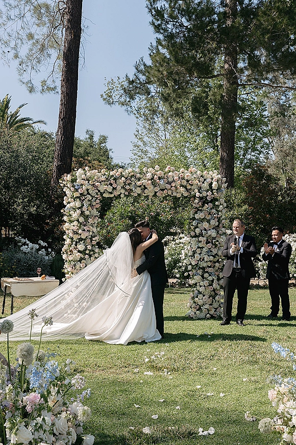 Ceremony kiss as bride and groom embrace under a full floral arch of white and blush roses, veil trailing on a sunny garden lawn