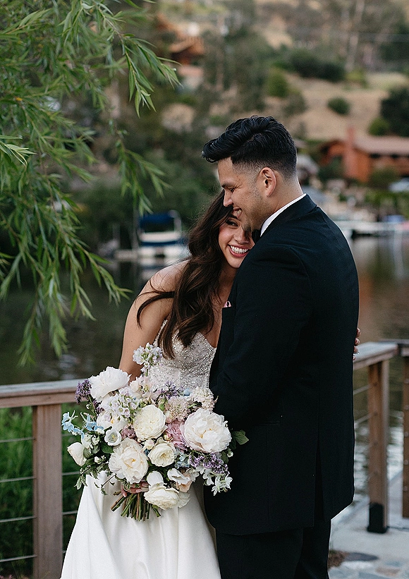 Couple portrait of bride and groom hug on a lakeside dock, bride in strapless gown holding a white and blush bouquet with lavender accents