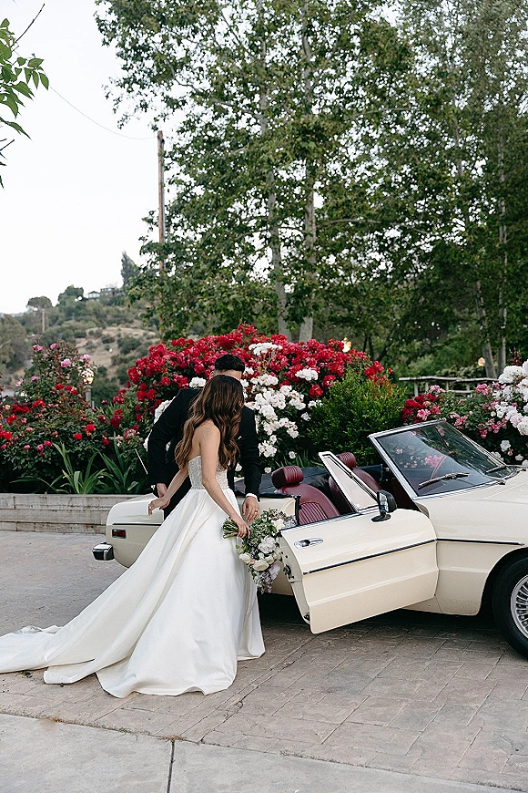 Wedding couple portrait of groom helping the bride step into a vintage convertible, her bouquet in hand beside rose bushes on a driveway