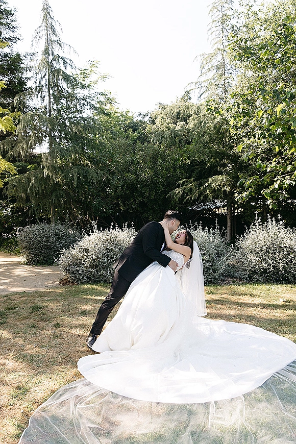 Wedding kiss portrait of the groom dipping the bride as her long veil and dress train flow in sunlight on a garden lawn