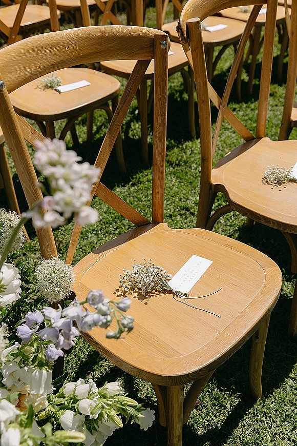 Ceremony seating with wood crossback chairs lined on a grass lawn, aisle flowers with baby’s breath accents and programs on chairs