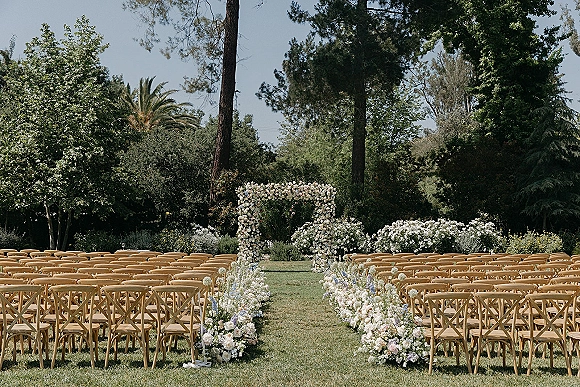 Ceremony setup for an outdoor wedding ceremony with a white floral arch and flower-lined aisle, wood chairs arranged on a garden lawn