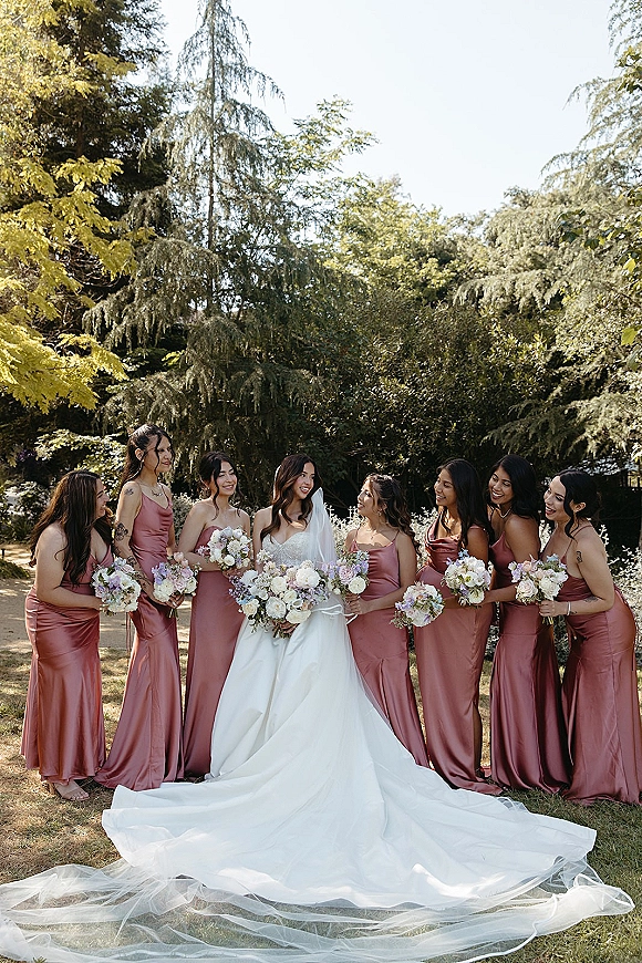 Bridesmaid group photo with bride and bridesmaids in satin dresses holding bouquets, bride in long veil on a green garden lawn in daylight