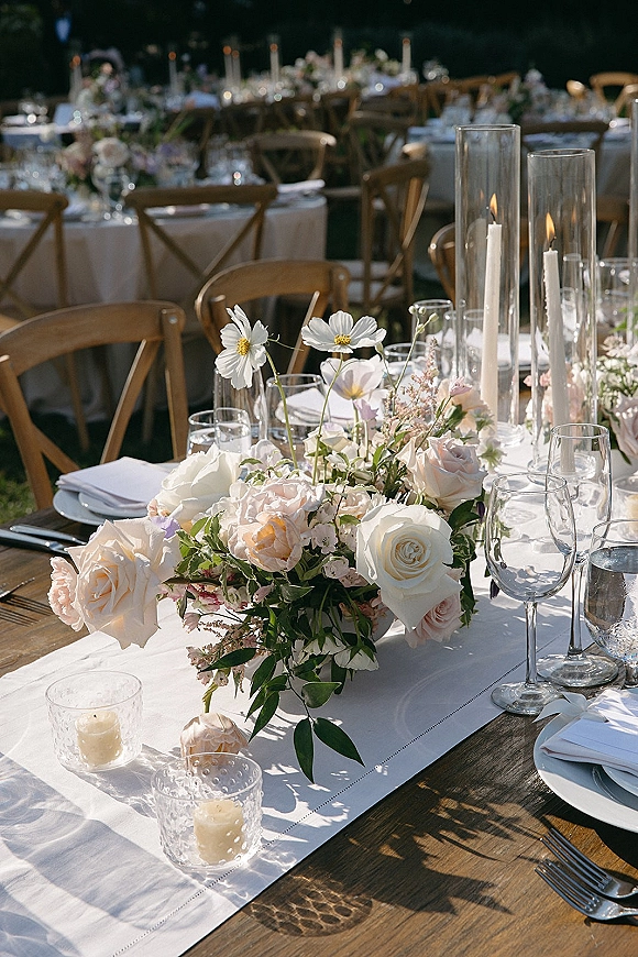 Reception tablescape with white table runner, rose and cosmos centerpiece, taper candles in glass hurricanes on round tables on a lawn