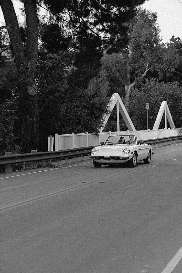 Wedding getaway car, a vintage convertible with greenery garland, driving over a bridge road bordered by guardrails and trees