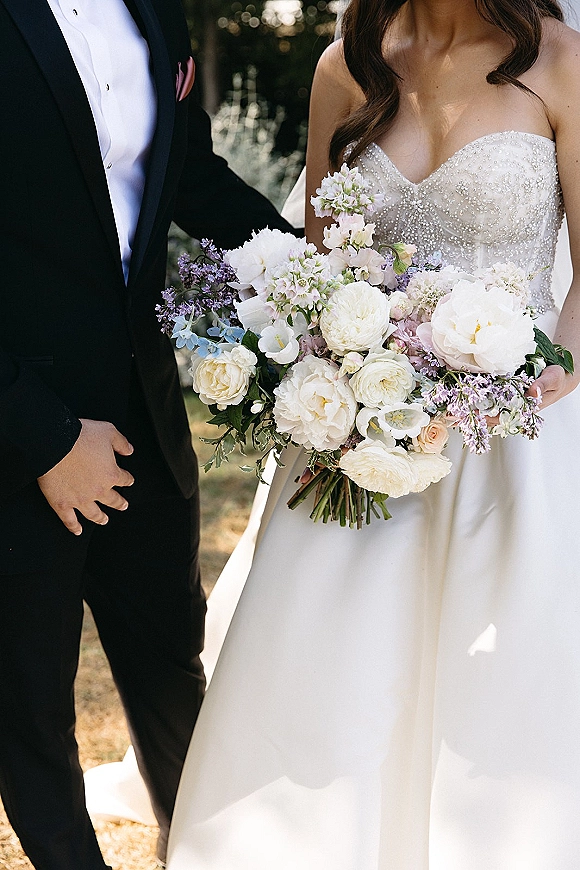 Couple portrait of bride holding a white and lavender bouquet beside groom in black tuxedo, in a sunlit garden with trees